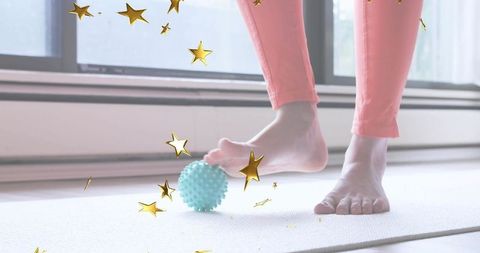Woman rolling foot over spiky massage ball on yoga mat near window for at-home foot therapy