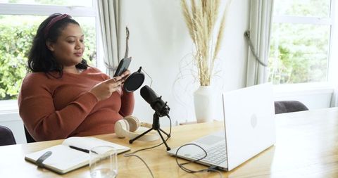 Woman preparing podcast episode at home desk with microphone, laptop and smartphone