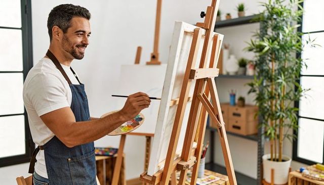 Smiling Male Artist Painting on Canvas in Bright Studio Wearing Denim Apron and Holding Palette