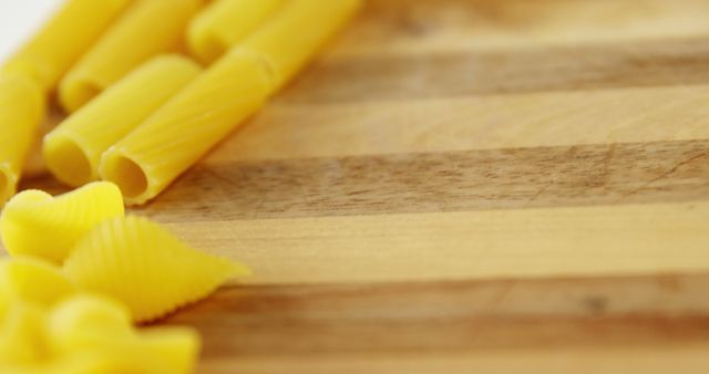 Uncooked pasta is arranged on a wooden cutting board, with copy space. Different pasta shapes like penne and conchiglie offer a variety of textures for culinary creations.