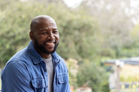 Smiling african american man leaning on porch rail wearing denim shirt and sweater in garden
