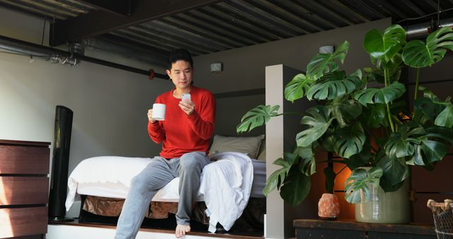 Man in casual attire sitting on bed in a modern loft apartment, holding coffee and smartphone. Ideal for themes related to modern living, technology, relaxation, and morning routines. Could be used in advertisements for lifestyle brands, interior design articles, or health and wellness content.