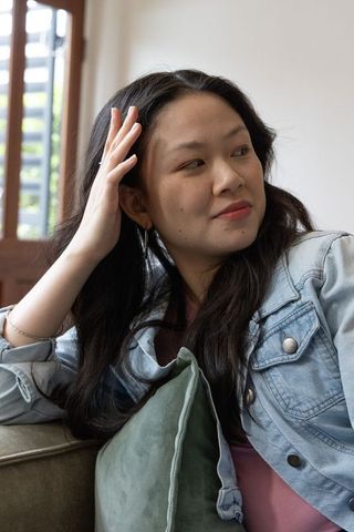 Asian woman relaxing on sofa touching hair, holding green cushion, wearing denim jacket