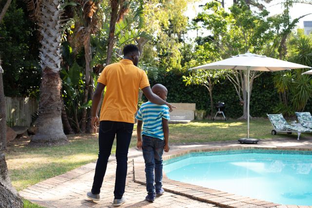 African American father and son walking in the backyard of their home, near the swimming pool, father pointing. Family and domestic life.