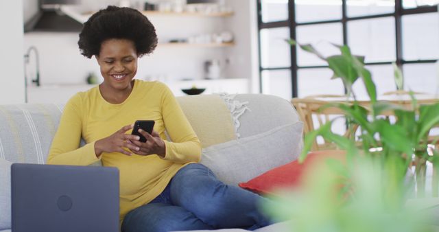 Happy african american woman sitting on sofa using laptop and smartphone. domestic lifestyle, spending free time at home.