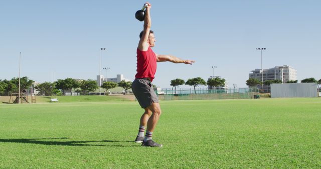 Fit caucasian man exercising outdoors, squatting and lifting kettlebell weight with one arm. cross training for fitness at a sports field.