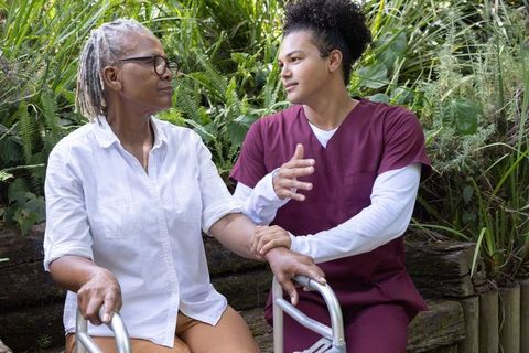 Kneeling caregiver supporting african american senior with walker in outdoor garden on bench