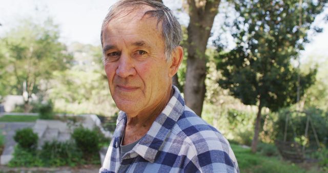 Elderly man wearing plaid shirt standing in a sunny park with lush greenery in the background. Suitable for content related to senior lifestyle, healthy aging, outdoor activities for the elderly, or nature walks for seniors.