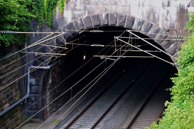 Deep railway tunnel entrance with stone archway and ivy-covered surroundings, leading into darkness, crisscrossed by electric wires above the train tracks. Suitable for use in articles about transportation infrastructure, travel, or industrial architecture. Can also be used for creating visuals for concepts like journey, exploration, or nature reclaiming man-made structures.