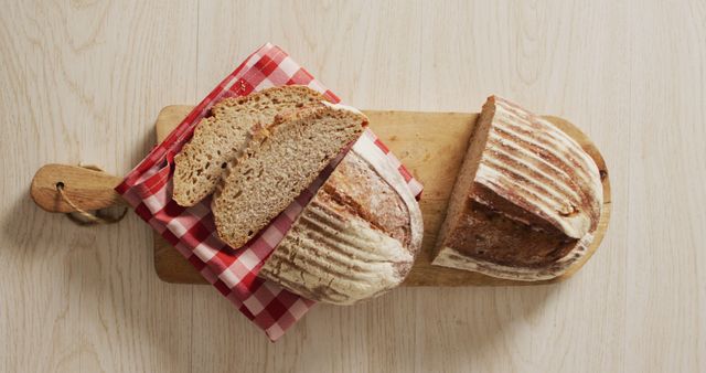 This photo features freshly baked sourdough bread with two slices placed on a wooden cutting board and a red and white checkered napkin. Ideal for use in culinary blogs, bakery websites, recipe books, or advertisements for bakeries showcasing artisanal breads. Perfect for promoting homemade and rustic baking themes.