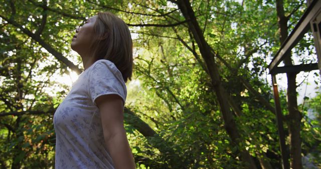 The image features a woman standing in a green forest, looking upward while sunlight filters through the trees. This conveys calmness, relaxation, and a connection with nature. It is suitable for themes such as mental health, meditation, nature retreats, environmental conservation, and outdoor activities promotions.