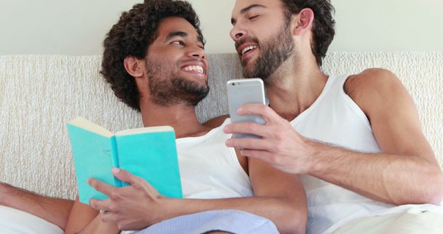 Two men are lying together in bed, one reading a book and the other holding a phone. They appear to be happy and affectionate, smiling at each other. The scene conveys a sense of intimacy, relaxation, and bonding. This image can be used to represent love, relationships, or an intimate moment in a home setting.