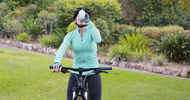 Woman cycling through a green park while hydrating, expressing outdoor fitness and a healthy lifestyle. Ideal for content related to fitness, outdoor activities, hydration tips, and wellness. Can be used in articles, blog posts, or promotional material for health and fitness products.