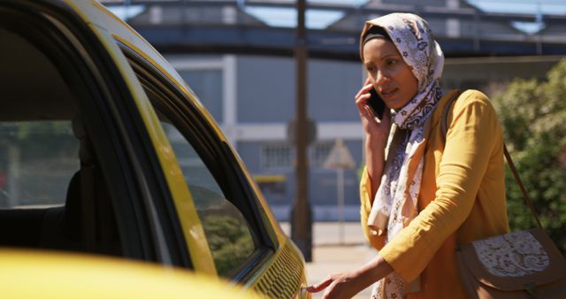 Front view of a young biracial woman wearing a hijab, talking on the smartphone and getting in a taxi in a city