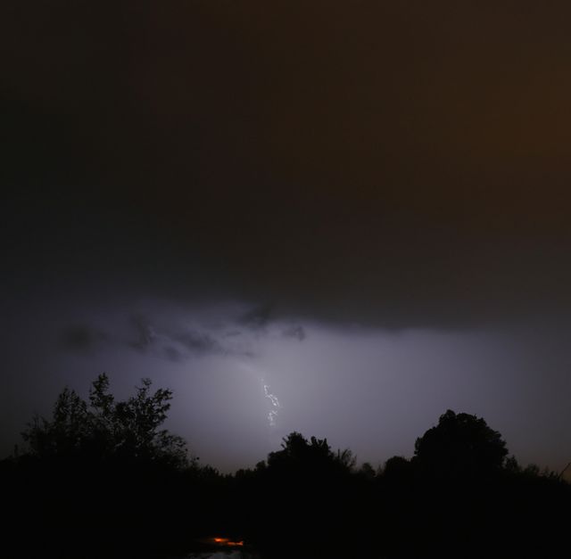 Capturing the dramatic ambiance of a nighttime thunderstorm with a visible bolt of lightning. Silhouetted trees add to the moody atmosphere. This can be used in weather-related articles, presentations on natural phenomena, or backgrounds for dramatic scenes.