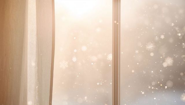 Golden morning snowfall through sheer curtain and window frame creating cozy winter glow