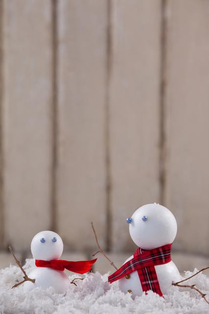 Two snowmen, one larger and one smaller, are adorned with scarves and surrounded by fake snow. The larger snowman wears a plaid scarf, while the smaller one has a red scarf. This image is perfect for holiday cards, winter-themed decorations, festive advertisements, and seasonal blog posts.