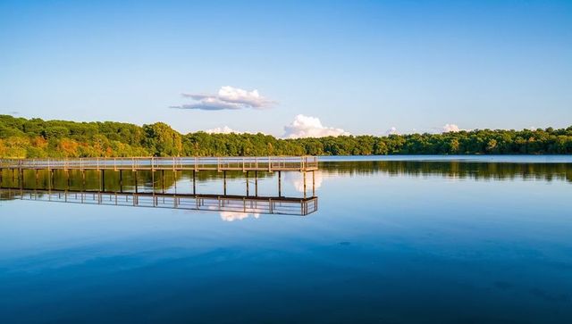 Extending pier with railings reflecting on glassy lake at golden hour with treeline