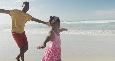 Running daughter skipping along shoreline while parent playing and laughing on sunny beach