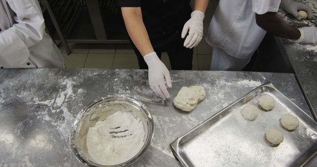 Bakers working together kneading dough and preparing raw buns in a professional kitchen setting. Useful for content related to baking, culinary education, professional kitchens, teamwork, and bakery-related services or products.