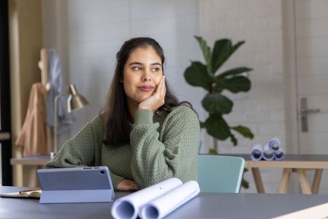 Asian woman working thoughtfully with tablet and rolled plans in minimalist home office