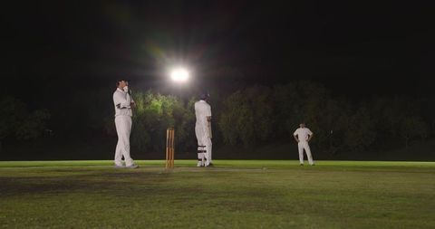 Night cricket match under floodlights with diverse players in whites at wicket on grass