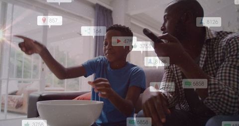 Father and son watching tv while pointing at screen with floating social media notifications