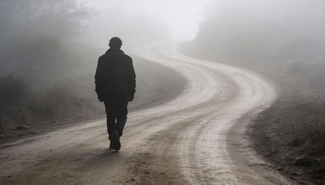 Walking man along foggy winding dirt road at dawn, solitary silhouette on misty path