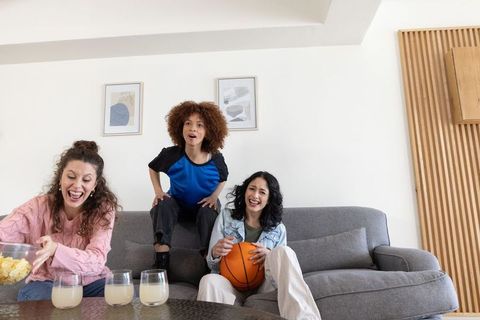 Friends watching game on couch sharing snacks and laughing with basketball