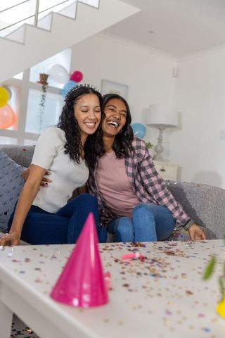 Female friends hugging and laughing on sofa celebrating birthday with colorful balloons