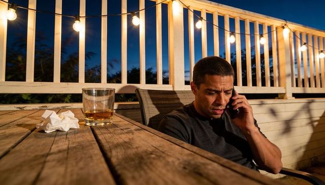 Man talking on phone at dusk on deck with whiskey glass and string lights, tense moment
