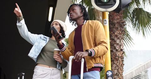 Diverse young travelers pointing way at urban transit curb with luggage, coffee, and phone