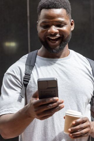 Smiling man checking smartphone while holding coffee and wearing backpack in urban commute