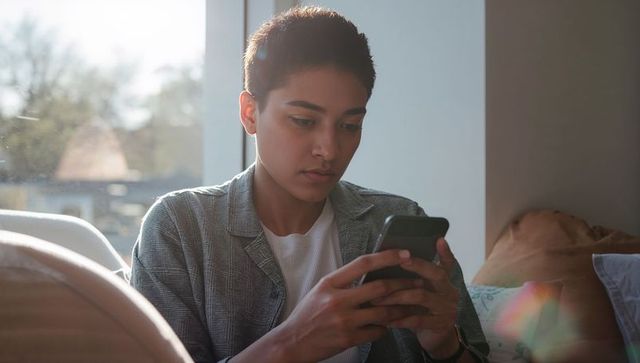Young woman holding smartphone checking messages on couch by sunlit window