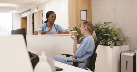 Two nurses taking coffee break and chatting at modern clinic reception with stethoscope