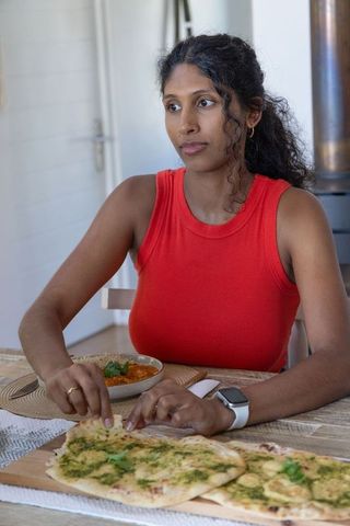 Woman reaching for herb-topped flatbread at cozy kitchen table with tomato dip and smartwatch