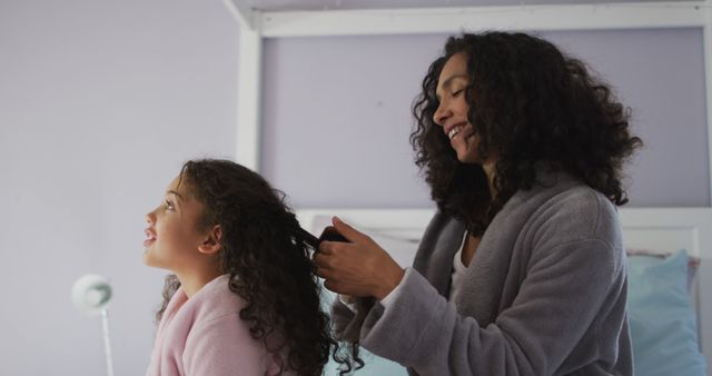 This heartwarming scene of a biracial mother and daughter brushing hair together in a cozy bedroom highlights the love and care in daily family routines. It can be used in parenting articles, family-focused advertisements, home decor features, and lifestyle blogs. The image evokes warmth, connection, and the importance of quality family time.