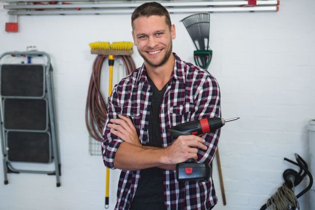 This image features a confident male carpenter holding a power drill in a workshop. He is smiling and wearing a plaid shirt, surrounded by various tools and equipment. This image can be used for promoting DIY projects, home improvement services, handyman services, or professional carpentry work.
