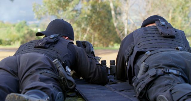 Rear view of two caucasian male soldiers in black lying in wait outdoors with binoculars and guns. Military services, weapon, war, army training and armed forces, unaltered.