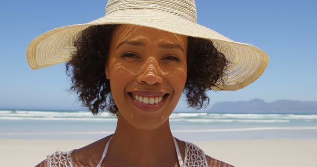 A smiling young African American woman wearing a sun hat enjoys a sunny day at the beach, with copy space. Her radiant smile and stylish hat capture the essence of a perfect summer getaway.