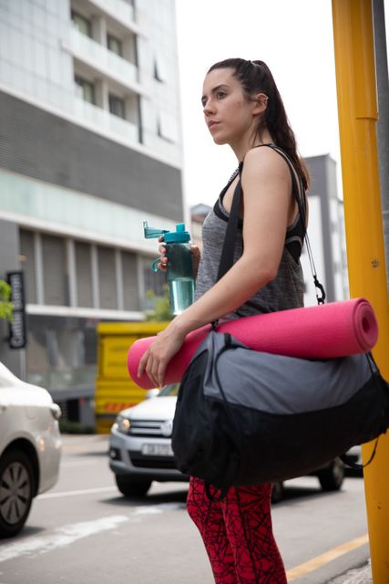 Fit woman walking in urban street carrying sports bag and yoga mat, holding water bottle. Ideal for promoting active lifestyle, fitness training, workout routines, and healthy living. Suitable for use in fitness blogs, health magazines, and exercise-related advertisements.