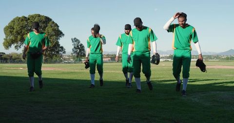 Baseball Team Walking Across Sunlit Field in Green Uniforms Carrying Gloves and Caps