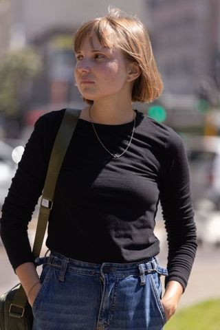 Young woman standing on city sidewalk looking thoughtful in black top and belted jeans