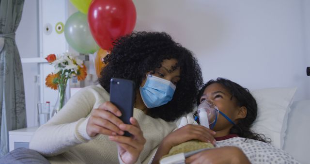 Mother taking a selfie with her sick daughter in hospital, both wearing masks, colorful balloons in background, flowers on bedside table, healthcare and recovery. Can be used for advertising family support, hospital care, healthcare services, parent-child relationships, medical insurance, hospital promotions.
