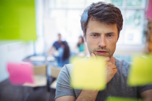 Male executive concentrating on sticky notes in an office environment. Ideal for depicting brainstorming sessions, strategic planning, and professional teamwork. Useful for business, productivity, and creative process themes.