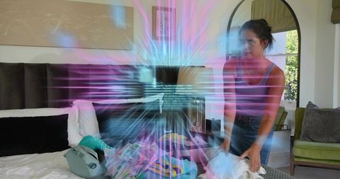 Young woman making bed and packing backpack with neon holographic light burst in bedroom