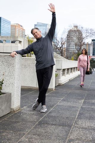 Middle-aged man stretching on urban promenade railing while woman walking with casual bag