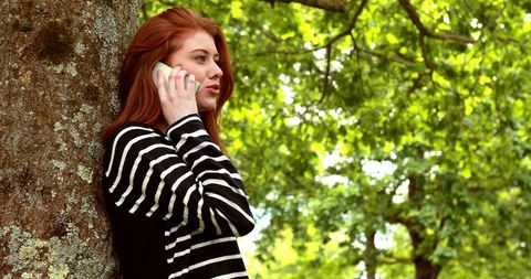 Redhead woman talking on phone by tree on sunny day