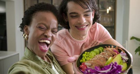 Diverse couple laughing while holding colorful poke bowl and taking selfie in home kitchen
