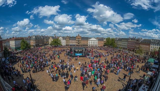 Vibrant crowd gathering around central stage in historic market square under blue sky